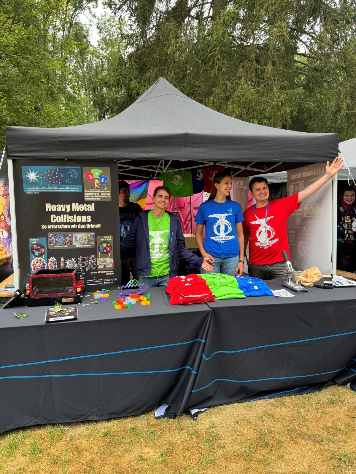 Drei Personen in farbigen T-Shirts in rot, grün und blau hinter einem Tisch mit Informationsmaterial.
