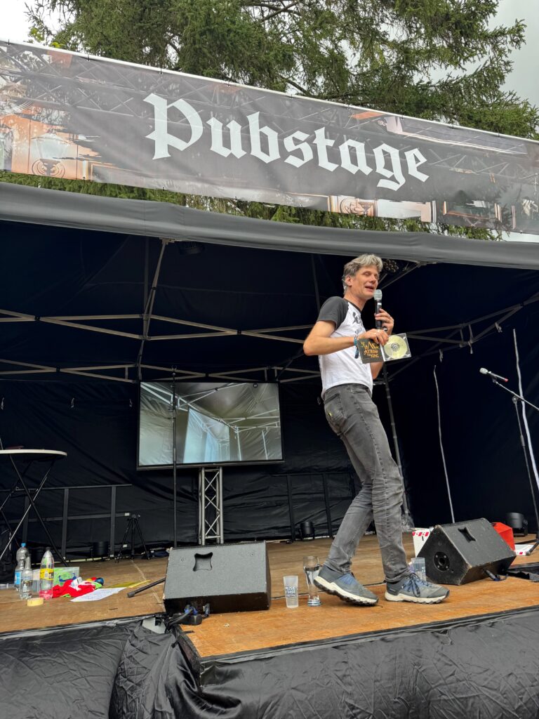 Ein Mann mit Mikrofon auf einer Bühne mit schwarzem Hintergrund. Er trägt eine schwarze Jeans und ein schwarz-weißes T-Shirt.