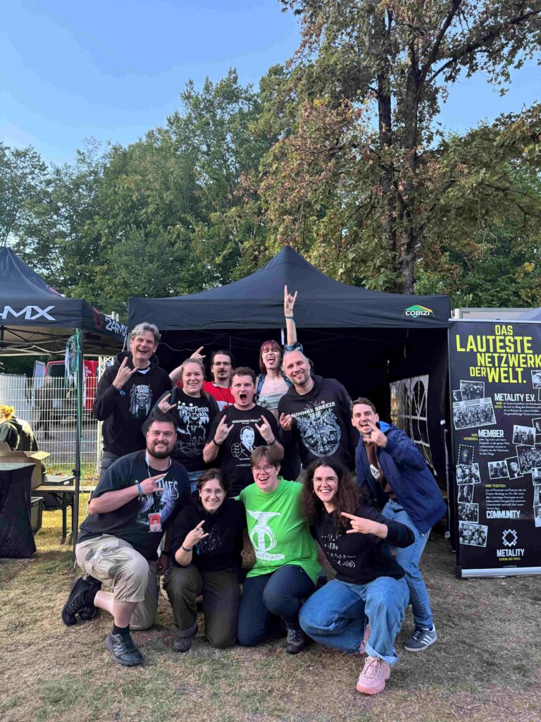Gruppenbild vor einem Pavillon. Die Personen tragen vor allem schwarze T-Shirts und zeigen lachend das Pommesgabel-Symbol (ausgestreckter Zeigefinger und kleiner Finger).